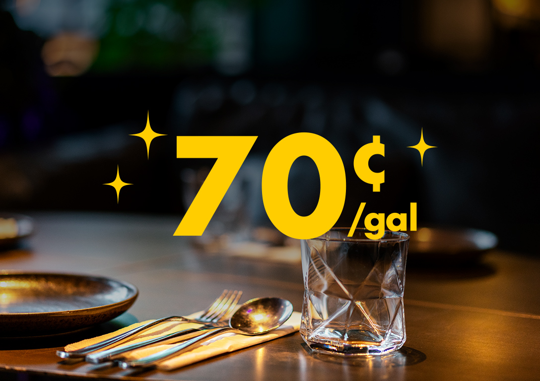 70¢ per gallon. A dimly lit restaurant table is set with plates, cutlery, napkins, and a glass of water, suggesting an evening out is now more affordable.