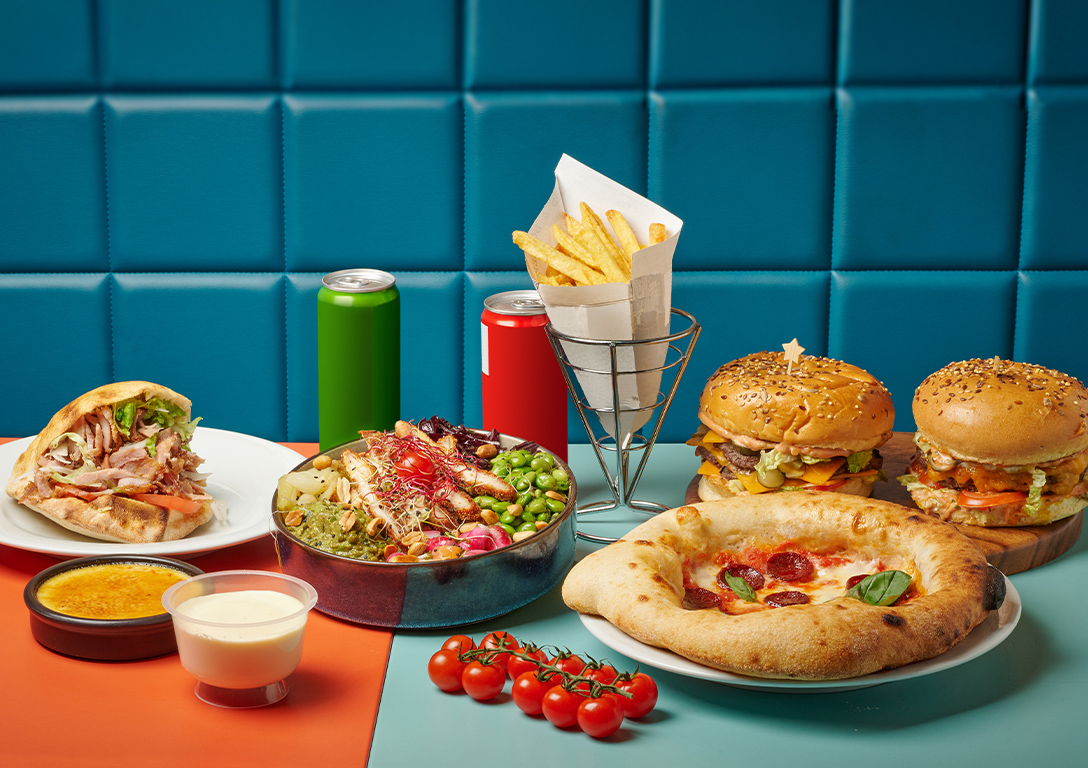 A colorful spread of fast food including burgers, pizza, a salad bowl, a kebab, fries, crème brûlée, and drinks, arranged on a two-toned table against a bright blue wall.